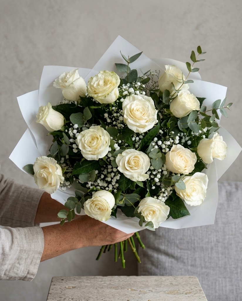 Ramo grande de rosas blancas y eucalipto con flores pequeñas, envuelto en papel, sostenido por manos.