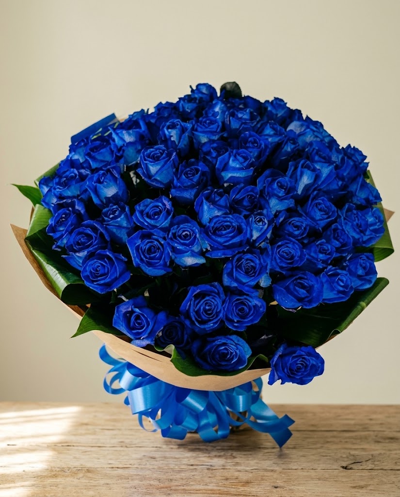 Ramo redondo de rosas azules con lazos azules y hojas verdes, sobre mesa de madera clara.