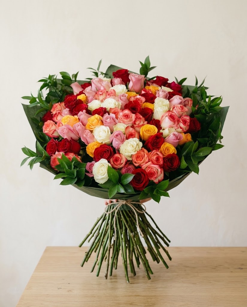 Ramo grande de rosas rojas, rosadas y amarillas con hojas verdes, atado con lazos sobre mesa.