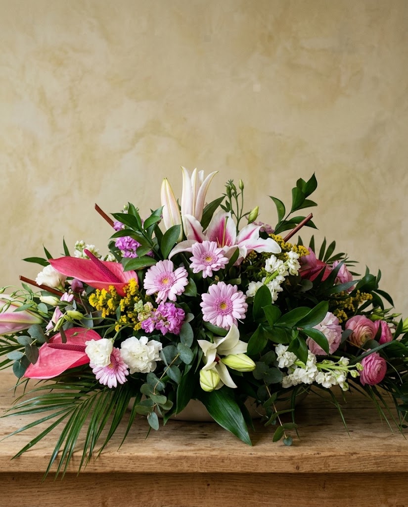 Arreglo floral multicolor con lirios, gerberas y flores pequeñas sobre tabla de madera, fondo beige.